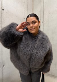 Manteau en fausse fourrure grise avec des manches oversize, doté d'un col montant et d'une texture douce, porté avec un pantalon gris foncé sur un fond en béton.