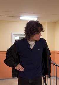 Tumma nahkatakki päällä tummansininen poolopaita, jossa pieni vihreä logo, kerroksittain useita kultaketjuja ja mustat housut.