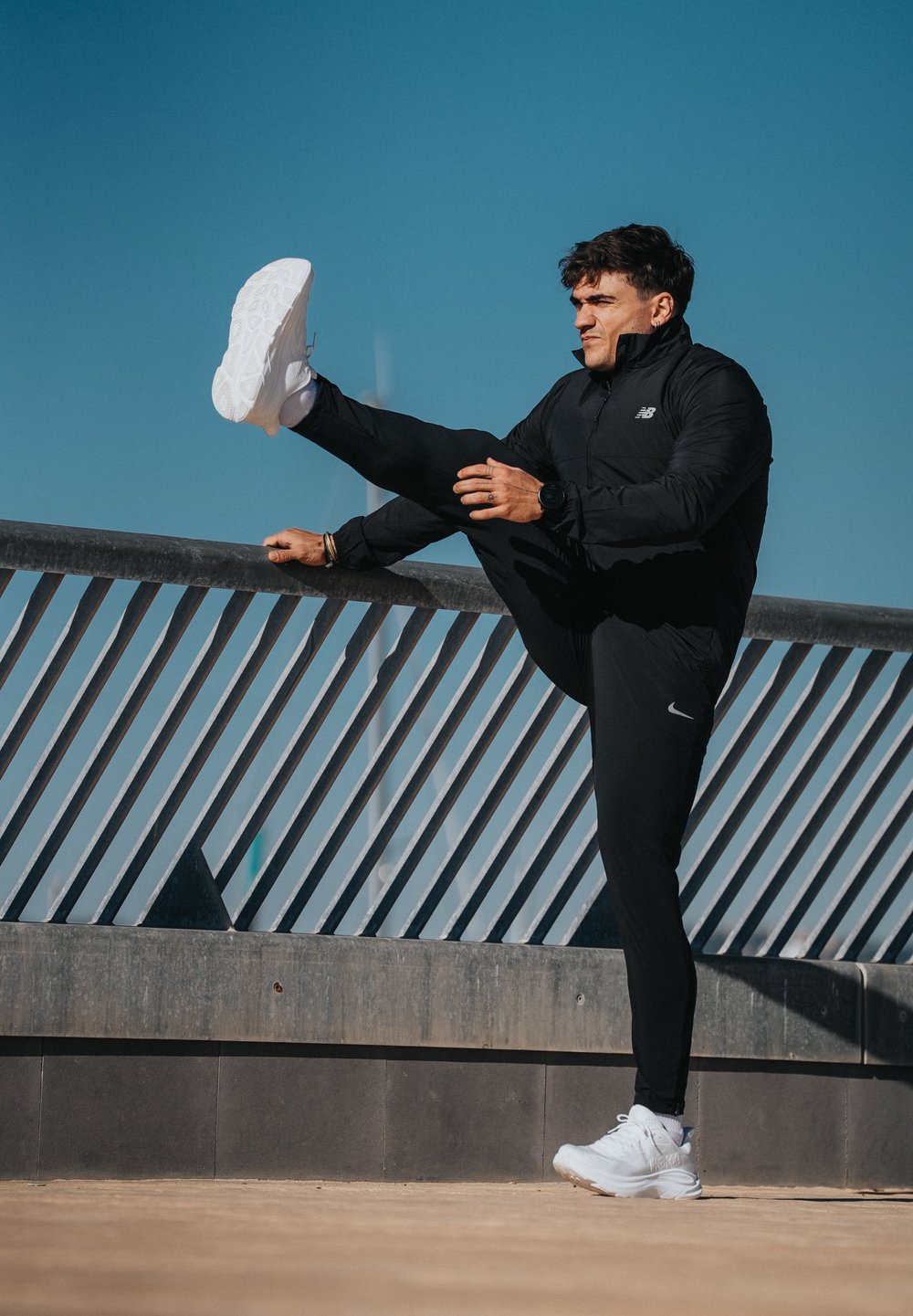 Czarna sportowa kurtka zapinana na zamek, dopasowane czarne spodnie i białe trampki. Osoba rozciąga nogę na poręczy pod bezchmurnym, błękitnym niebem.