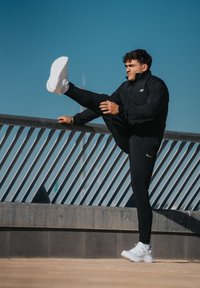 Czarna sportowa kurtka zapinana na zamek, dopasowane czarne spodnie i białe trampki. Osoba rozciąga nogę na poręczy pod bezchmurnym, błękitnym niebem.