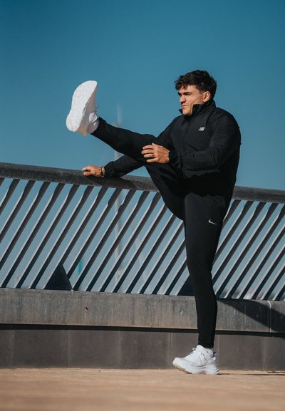 Chaqueta deportiva negra con cremallera, pantalones negros ajustados y zapatillas blancas. Persona estira la pierna apoyándose en una barandilla bajo un cielo azul despejado.