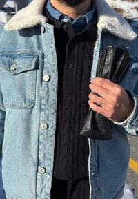 Veste en jean avec col en shearling, pull noir en laine torsadée en dessous, et gants en cuir noir tenus à la main. Textures nettes et couleurs contrastées.