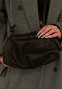 Čierna kožená kabelka s naťahovaným dizajnom, hladkou textúrou a zapínaním na zips, držená v ruke s dlhými, zložitými nechtami.