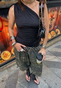 Črna enostranska majica, široke hlače z vojaškim vzorcem, srebrni zakoviki na pasu, črna mini torbica s verižnim trakcem, drži pijačo.