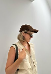 Brun kasket med flossede kanter, lavet af bomuldsstof, iført med en beige ærmeløs vest og en mørkegrøn taske. Tekstureret overflade og afslappet design.