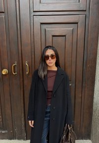 Manteau en laine noire avec grands boutons sur un haut bordeaux ; jean en denim foncé ; sac à main marron ; lunettes de soleil marron ; arrière-plan en panneaux de bois.