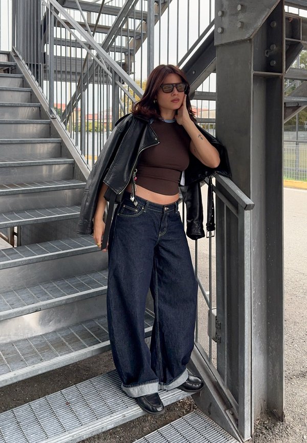 Femme portant des lunettes de soleil, une veste en cuir noire posée sur les épaules, un crop top et un jean large debout sur un escalier en métal à l'extérieur.