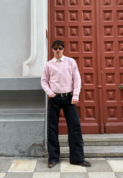 Hombre con cabello rizado y gafas de sol, vestido con un suéter rosa y pantalones negros anchos, de pie frente a una puerta roja con paneles y una pared de piedra.