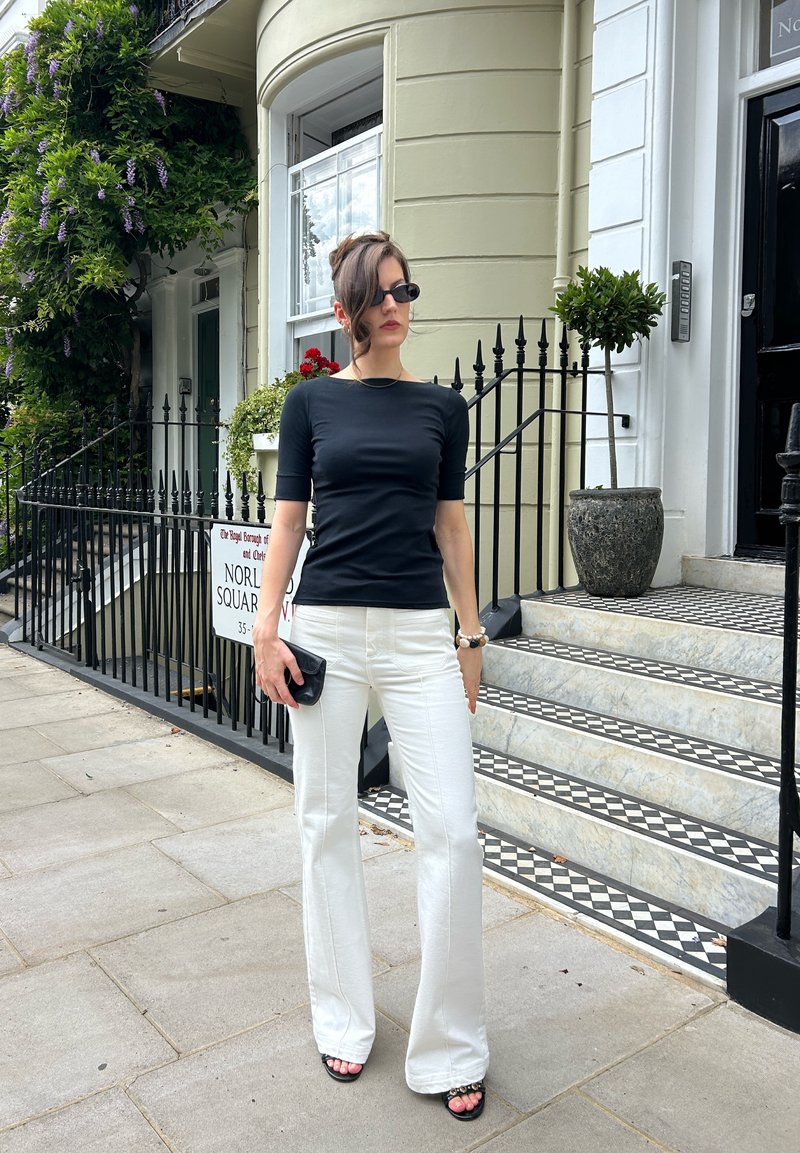 Femme en haut noir et pantalon blanc debout sur le trottoir à côté d'une maison de ville avec des rampes noires et des marches carrelées avec motifs.