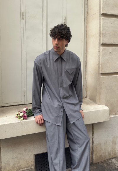 Joven con cabello rizado que lleva una camisa y pantalones grises, sentado en un borde con un pequeño ramo de flores a su lado.