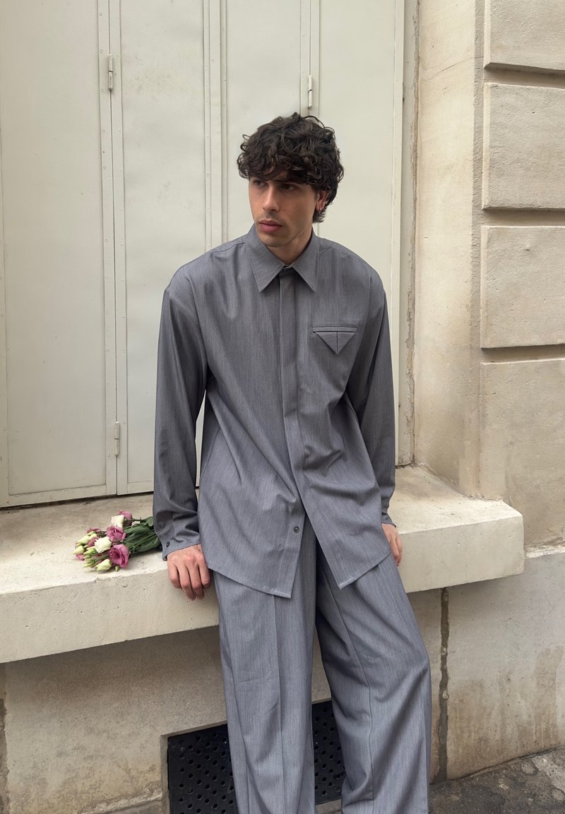 Young man with curly hair wearing a gray shirt and pants, sitting on a ledge with a small bouquet of flowers beside him.