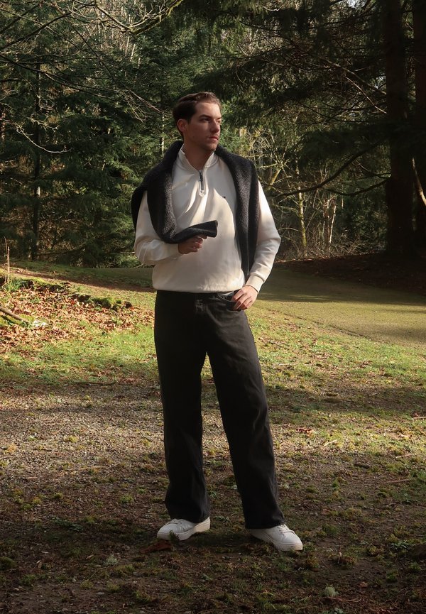 Άνδρας με λευκό πουλόβερ και μαύρο παντελόνι στέκεται σε μονοπάτι δάσους με ένα σκούρο πουλόβερ ριγμένο στους ώμους, κοιτάζοντας προς τα πλάγια.