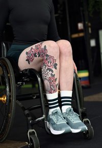 Personne assise en fauteuil roulant montrant une jambe tatouée avec des roses rouges et une illustration de femme, portant des chaussettes rayées et des chaussures de sport bleu sarcelle.