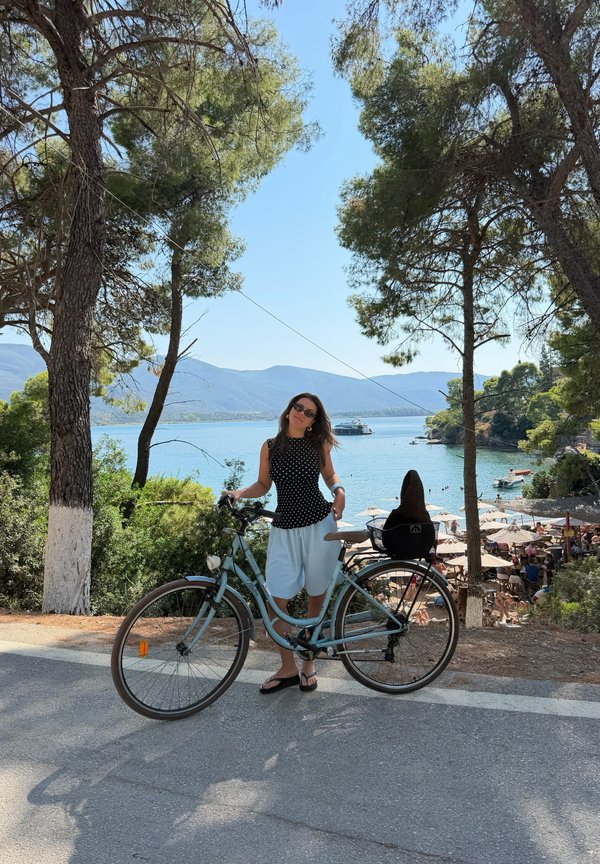 Femme debout avec un vélo sur une route ombragée donnant sur une plage ensoleillée avec des parasols, des arbres et un bateau sur une eau calme.