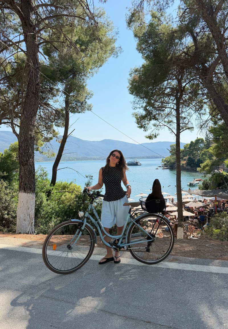 Γυναίκα στέκεται με ποδήλατο σε μια σκιερή οδό με θέα σε μια ηλιόλουστη παραλία με ομπρέλες, δέντρα και μια βάρκα σε ήρεμα νερά.