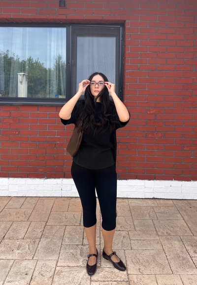 Parte superior negra, leggings negras, bolso marrón; cabello largo y oscuro. De pie sobre un suelo de baldosas frente a una pared de ladrillo rojo con una ventana.