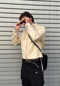 Béžová kožená bunda s vysokým límcem a zipem, nošená přes černé cargo kalhoty. Černý opasek s kovovými doplňky a malá černá taška.