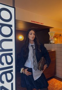 Giacca di jeans sopra una camicia bianca con una cravatta a quadri e una gonna nera. Tiene una borsa marrone, posando vicino a un'insegna illuminata e a luci decorative.