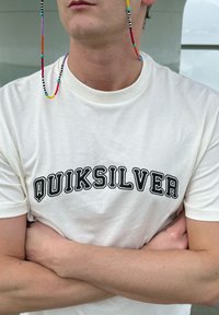 Άτομο που φοράει λευκό T-shirt Quiksilver με σταυρωμένα χέρια, πολύχρωμη αλυσίδα γυαλιώ ν με χάντρες κρεμασμένη από τα αυτιά, ουδέτερο φόντο.
