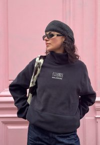 Femme portant un sweat Salomon noir, un béret noir à motif, des lunettes de soleil et un sac à épaule en fourrure noir et blanc, posant devant un mur rose.