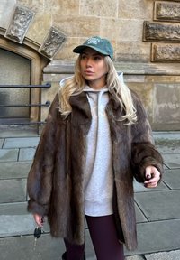 Manteau en fourrure brune à la texture douce, porté par-dessus un sweat à capuche gris. La tenue comprend une casquette de baseball verte et des leggings foncés. Des clés tenues dans une main.