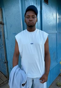 Balts bezgaruma krekls ar zilu "HUGO" logotipu, turēdams gaiši pelēku kapuci. Fons veidots no zila koka.