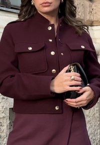 Burgunderfarbene, kurze Jacke mit goldenen Zubehörteilen, ausgestattet mit zwei Brusttaschen und einem Stehkragen. Die Person hält eine schwarze Clutch.