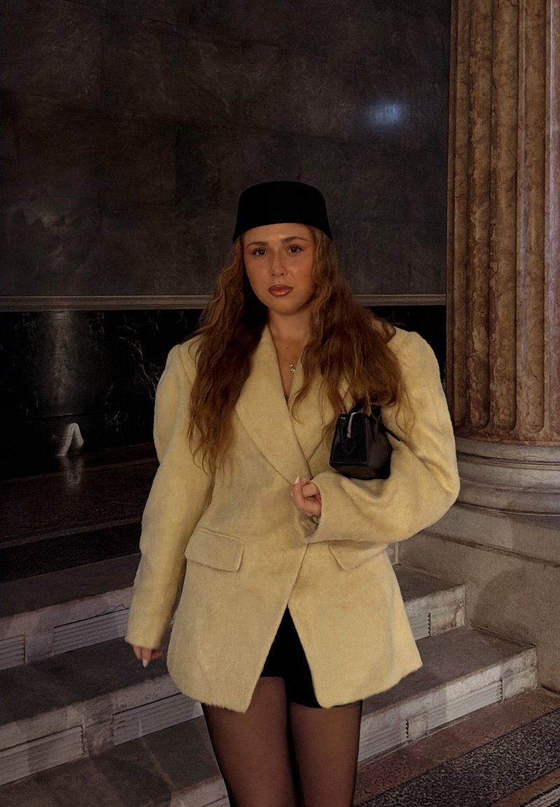 Femme aux cheveux longs et ondulés portant un chapeau noir, un manteau beige surdimensionné, un short noir, et tenant un petit sac à main noir, debout près de marches en marbre.