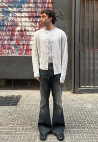 Ανοιχτό γκρι πλεκτό καράτιν με κουμπί, φορεμένο πάνω από γκρι πουκάμισο και σκούρο, φαρδύ παντελόνι, συμπληρωμένο με μαύρα παπούτσια.