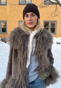 Jeune femme portant un bonnet en tricot foncé, un manteau épais en fausse fourrure grise, une écharpe claire et des gants, debout dehors dans la neige près d'un bâtiment jaune avec des fenêtres.