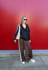 Jachetă neagră cu fermoar, cu dungi albe, pantaloni largi cu imprimeu de leopard, adidași albi și o geantă de umăr crem, toate pe un fundal roșu.