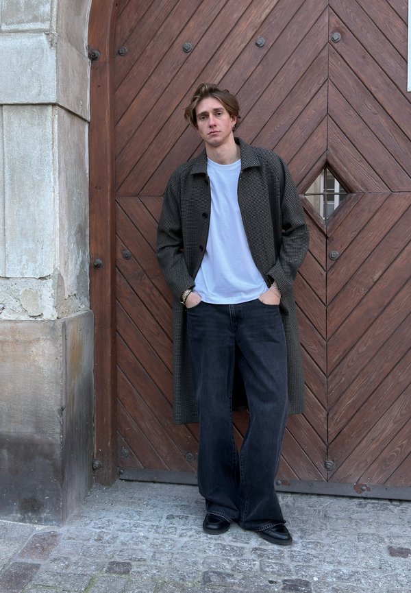Long, dark checkered coat over white T-shirt, paired with wide-leg black jeans and black shoes. Background features a wooden door with geometric patterns.