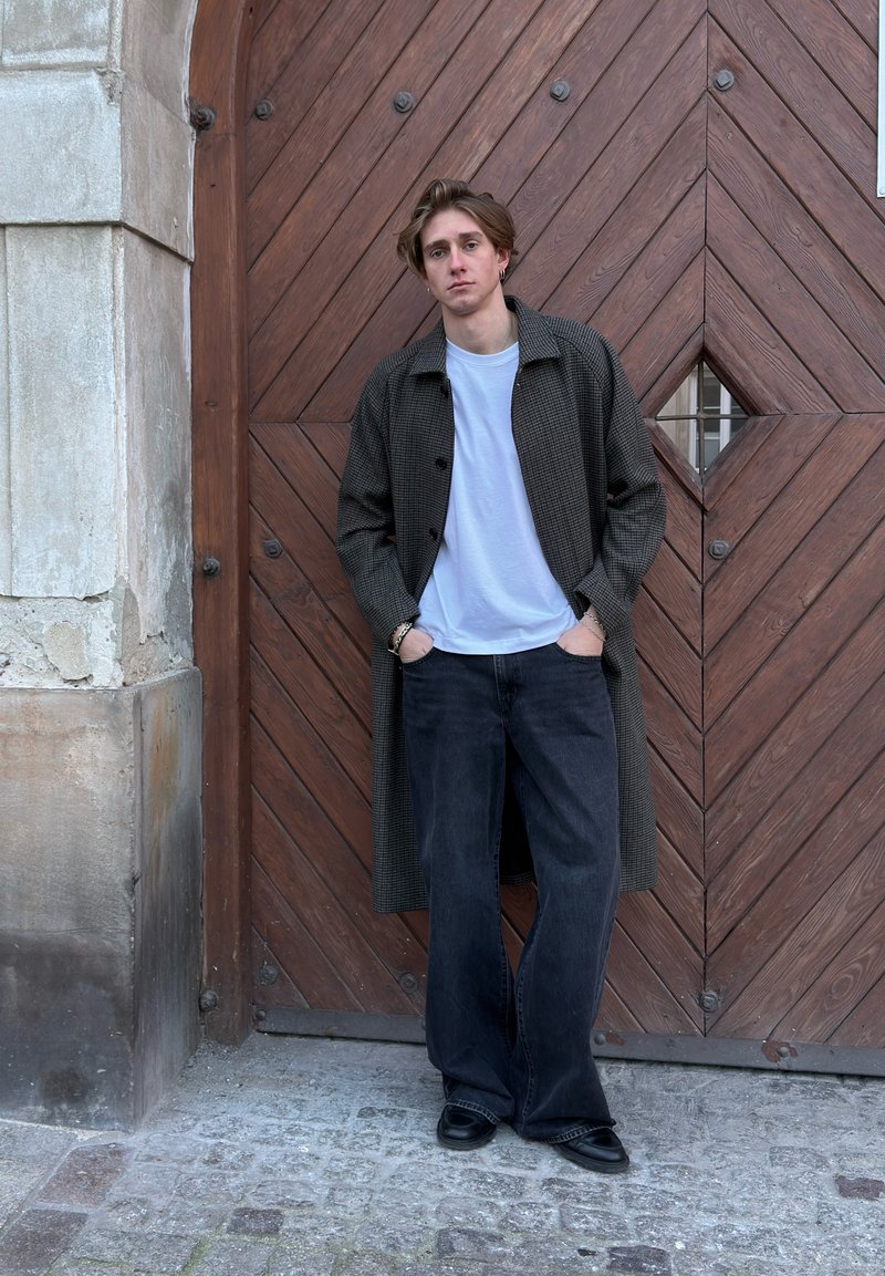 Lange, donker geruite jas over een wit T-shirt, gecombineerd met zwarte wijde jeans en zwarte schoenen. Op de achtergrond is een houten deur met geometrische patronen te zien.