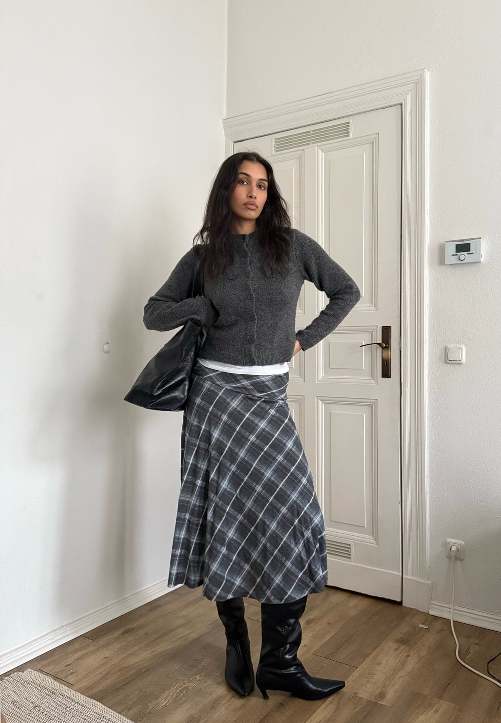 Femme debout à l'intérieur près d'une porte blanche, portant un cardigan gris foncé, une jupe écossaise grise, des bottes noires et portant un grand sac noir.