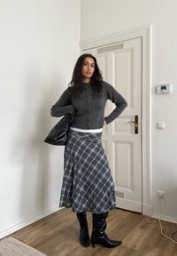 Femme debout à l'intérieur près d'une porte blanche, portant un cardigan gris foncé, une jupe écossaise grise, des bottes noires et portant un grand sac noir.