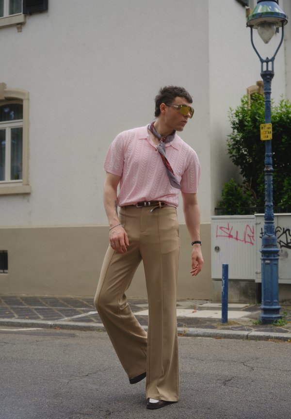 Mann in pinkem kurzärmligem Strickshirt, beigen weiten Hosen und einem um den Hals gebundenen Schal geht auf einer Straße in der Stadt nahe einer Straßenlaterne.