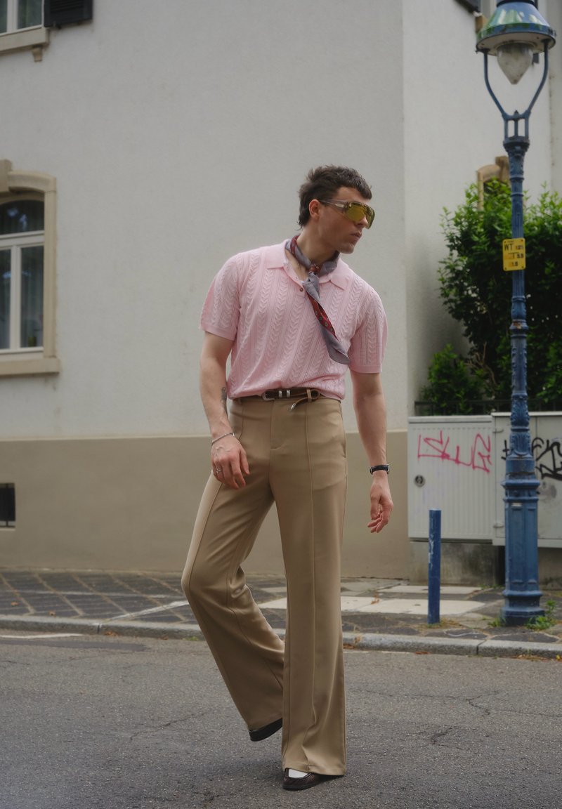 Mann in pinkem kurzärmligem Strickshirt, beigen weiten Hosen und einem um den Hals gebundenen Schal geht auf einer Straße in der Stadt nahe einer Straßenlaterne.
