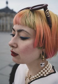 Fargerikt, kort hår med rosa, oransje og grønne strå. Har på seg brune solbriller, gulløredobber og et perlearmbånd. Blå eyeliner fremhever øynene.