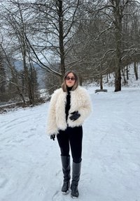 Femme portant une veste blanche en fourrure, une tenue noire, des gants et des bottes, debout sur un sol enneigé avec des arbres nus et un banc en arrière-plan.