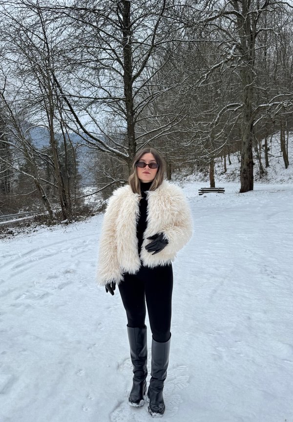 Žena v bielom chlpatom kabáte, čiernom oblečení, rukaviciach a čižmách stojí na zasneženom povrchu, v pozadí sú holé stromy a lavička.