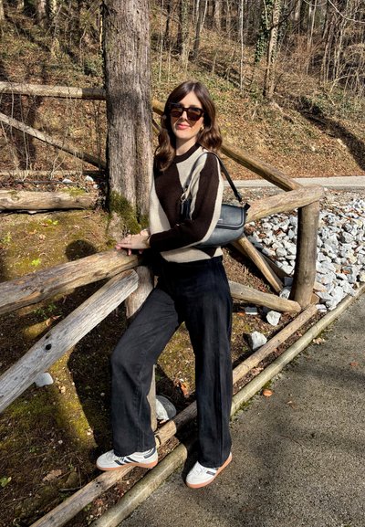 Jersey de punto con diseño geométrico en marrón y crema, combinado con pantalones de pierna ancha negros y zapatillas blancas. Los accesorios incluyen gafas de sol y un bolso negro.
