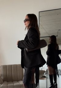 Femme en blazer noir, jupe en dentelle, bottes montantes et lunettes de soleil, portant un sac à bandoulière, debout près d'un canapé beige et d'un miroir.