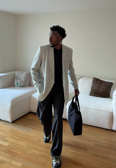 Hombre con blazer claro, pantalones y zapatos negros, llevando una bolsa negra, caminando en la sala de estar con sofá seccional blanco y cojín de estampado de leopardo.