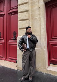 Pruun nahkjakk valge särgi kohal, pruunid laia säärega püksid, hoides pruuni kotti, seismas punaste uste lähedal, tekstureeritud kiviseina taust.