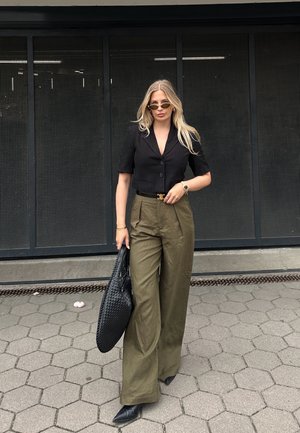 Blazer noir court, pantalon olive taille haute à jambes larges, sac à main tissé noir, escarpins noirs à talons pointus, lunettes de soleil rondes, cheveux texturés légers.