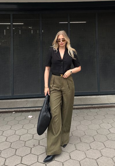 Blazer corto negro, pantalones de pierna ancha de talle alto en color oliva, bolso de mano tejido negro, tacones negros de punta, gafas de sol redondas, cabello claro y texturizado.