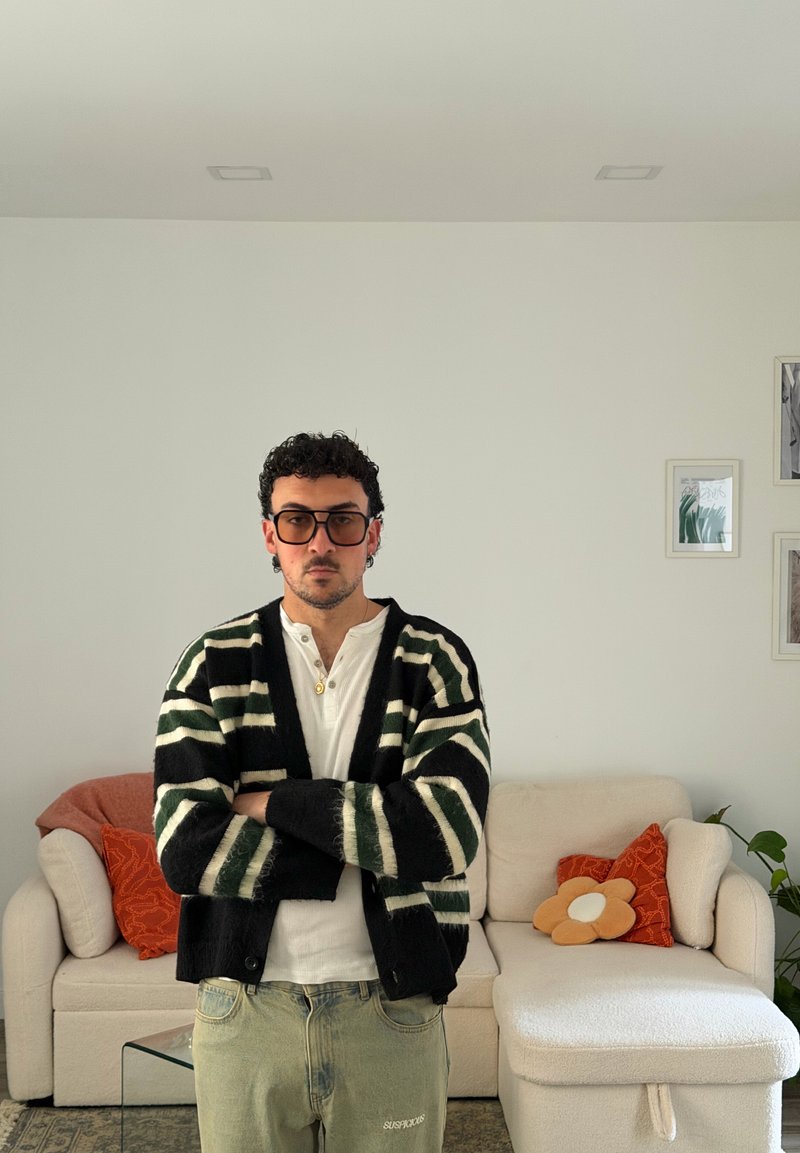 Homem com cabelo encaracolado e óculos, vestindo um casaco às riscas, está de braços cruzados numa sala de estar com um sofá branco e almofadas decorativas.
