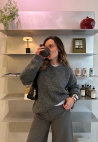 Γυναίκα με γκρι πουλόβερ που πίνει από μαύρη κούπα, στέκεται μπροστά σε ράφια με βιβλία, κονσέρβες, μπουκάλια και ένα φωτιστικό.