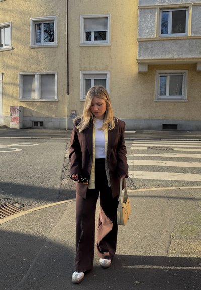 Joven mujer con traje marrón oscuro y zapatillas blancas, llevando un bolso beige y gafas de sol, caminando por una acera de la ciudad cerca de un paso de peatones.