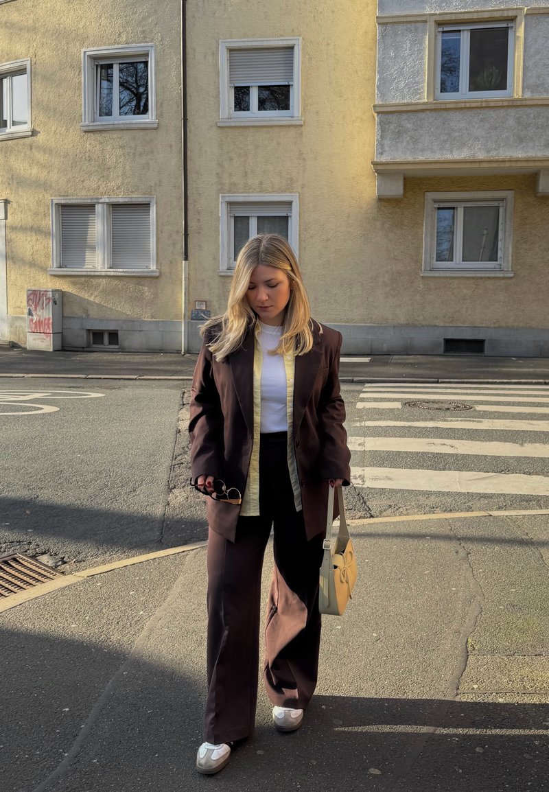 Jeune femme portant un costume brun foncé et des baskets blanches, tenant un sac à main beige et des lunettes de soleil, marchant sur un trottoir en ville près d'un passage pour piétons.